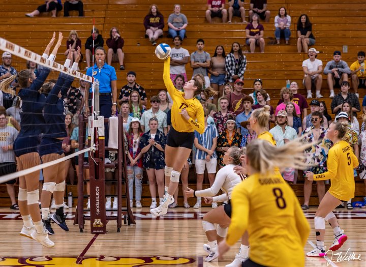 A volleyball player in a yellow jersey leaps to spike the ball over the net, while the opposing team sets up a block. Teammates prepare for the next move as spectators fill the bleachers in the background, watching intently. The scene is filled with energy as the teams compete on the court.